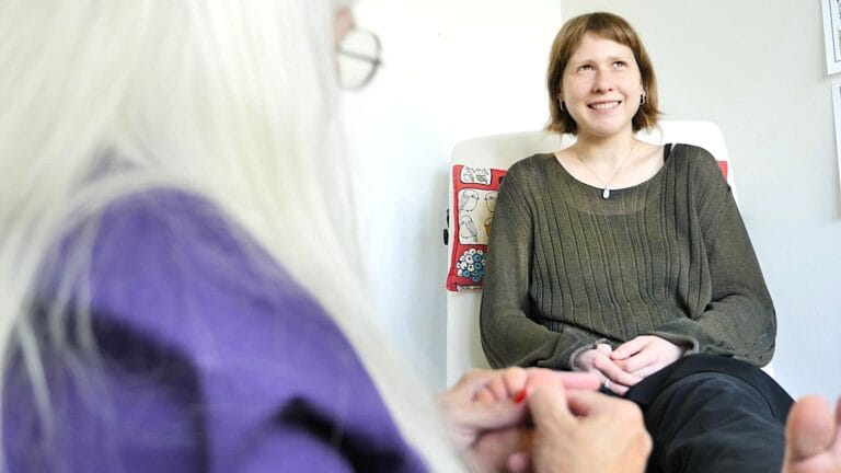 Nuori nainen istuu tuolissa ja hymyilee kun vyöhyketerapeutti painaa hänen jalkojaan.
